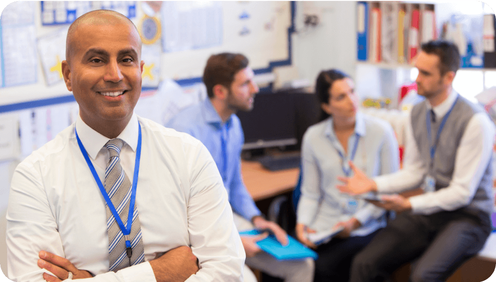 A confident school administrator with crossed arms smiling at the camera, with a background of educators in a collaborative meeting discussing school tech solutions.