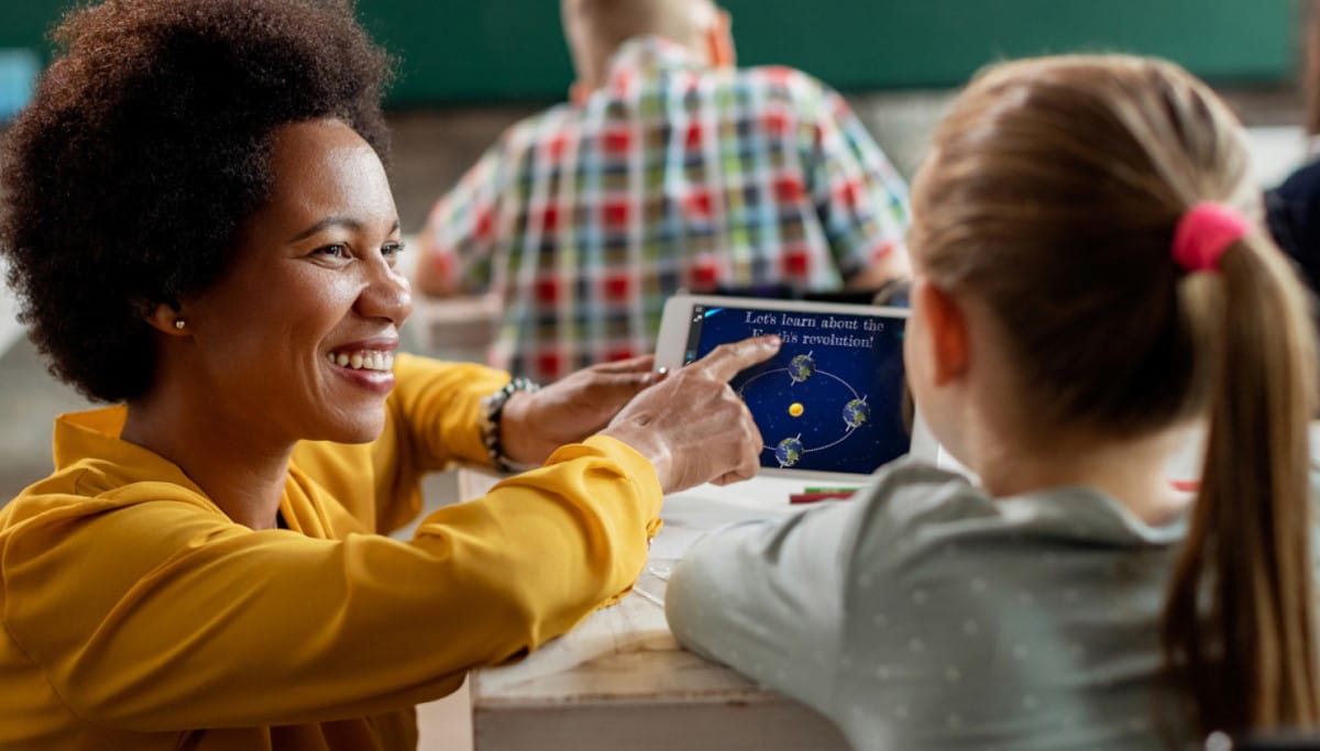 A teacher in a yellow blouse, using Lumio on a tablet, engaging a student with an interactive lesson about the Earth's revolution in a classroom setting.
