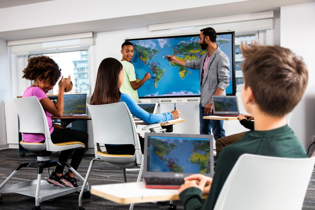 Students in a classroom look on as a teacher explains a topic on a SMART board with a world map.