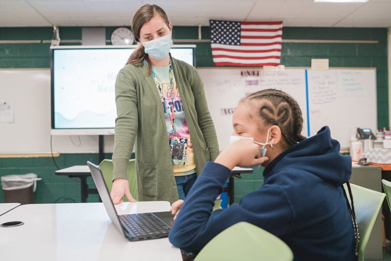 A teacher in a green cardigan leans over a student’s desk, assisting with a laptop in a classroom. The SMART Board displays a lesson in the background, accompanied by an American flag on the wall.